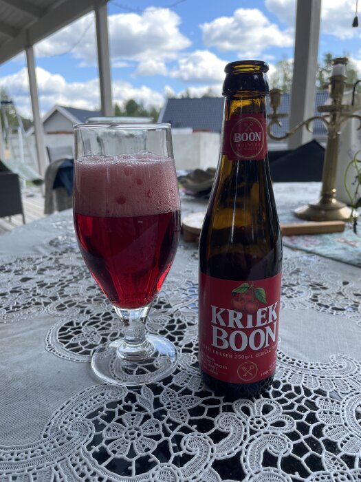 Ett glas med röd dryck och en Boon Kriek-flaska på spetsduk, utomhus med dagsljus och naturbakgrund.