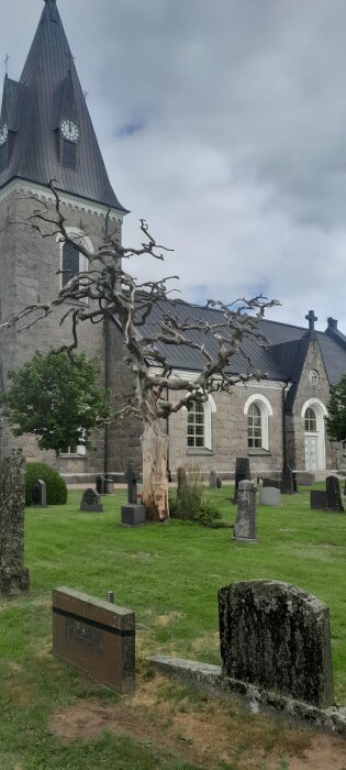 Kyrkogård med kala träd, gravstenar och kyrka i bakgrunden under mulet himmel.