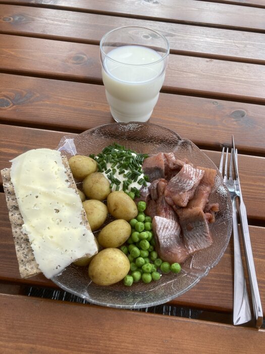 Ett måltid med knäckebröd, ost, potatisar, ärtor, sill, gräslök och ett glas mjölk på ett träbord.