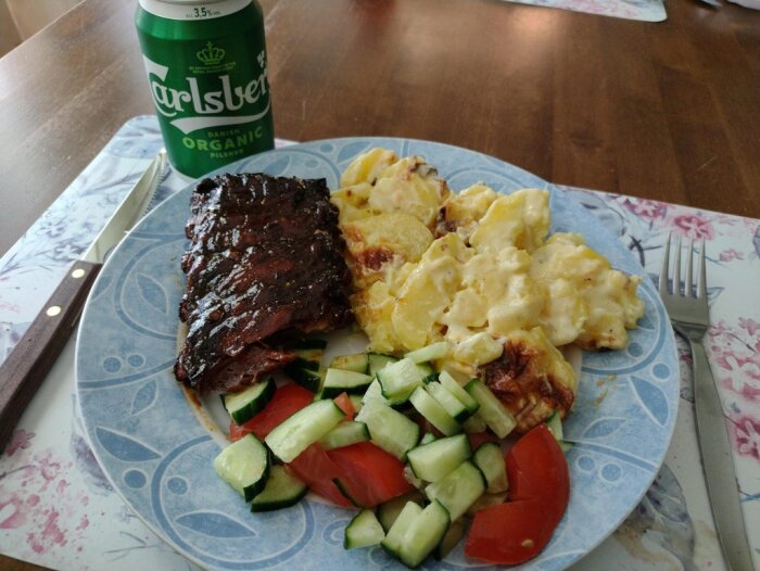 Grillat revbensspjäll, potatisgratäng, sallad, burköl Carlsberg, bestick, blommig tallrik, träbord.