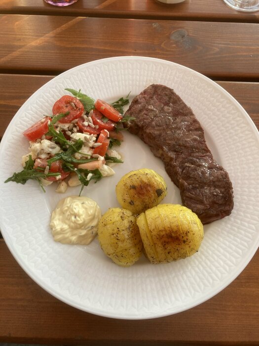 Grillad biff, klyftpotatis, sallad med tomat och fetaost, bearnaisesås på vit tallrik, träbord.