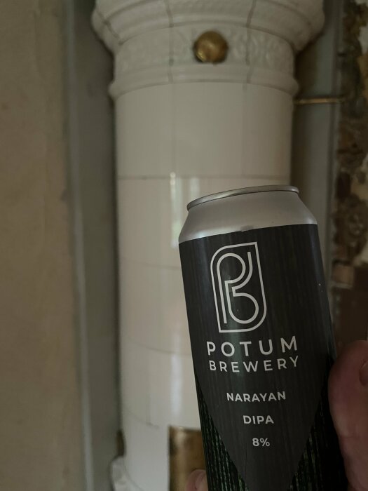 En hand håller en ölburk från Potum Brewery med texten "Narayan DIPA 8%" framför gammal kakelugn.