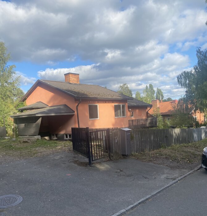 Ett orange tegelhus med carport, trädgårdsstaket och delvis molnig himmel.