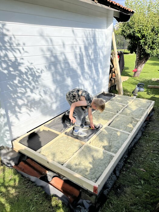 Barn hjälper till med byggnadsarbete utomhus, lägger isolering på trädäck under soligt väder.