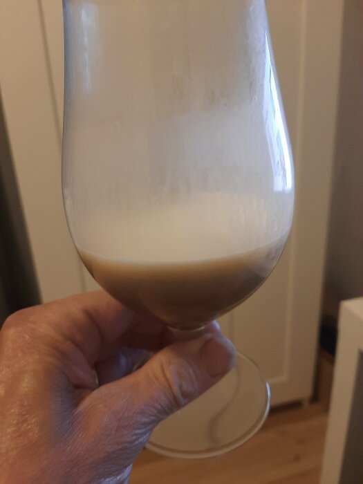 Hand håller ett glas med grumlig, skummande dryck, möjligen öl eller fermenterad dryck, inomhus.