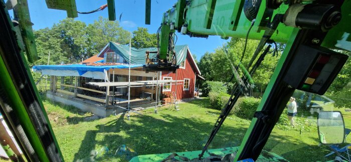 Vy från grävmaskinhytt, hus under renovering, person, grönska och stegar.