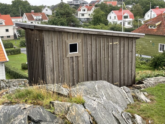 Träskjul med fönster på klippa, omgivet av grönska, med hustak i bakgrunden. Grått molnigt väder.