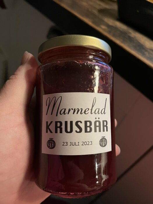 Ett burk hålls i en hand, etikett visar "Marmelad KRUSBÄR", daterad "23 JULI 2023".