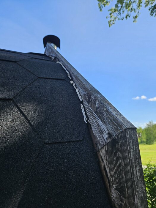 Närbild på mörkt takmaterial och trädetalj mot en klarblå himmel och trädgren.