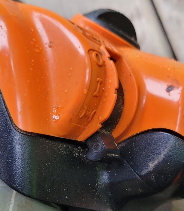 Närbild av en orange och svart plastdetalj, möjligtvis en del av en leksak eller verktyg, med vattendroppar och smuts.