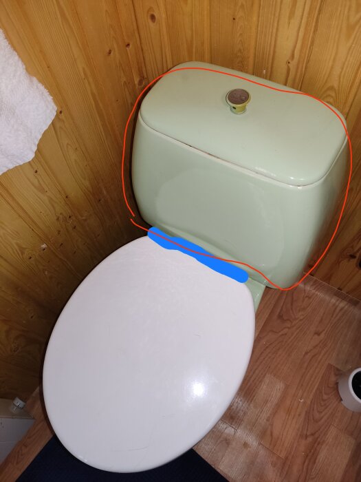 Toalettstol med öppet lock, träpanelvägg, grön spoltank, golv synligt.