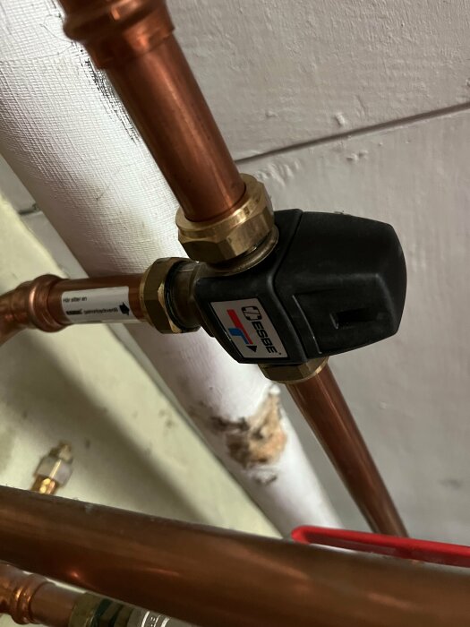 Kopparledningar med en svart ventil eller termostat, etiketter, teknisk installation, mot en vit vägg.