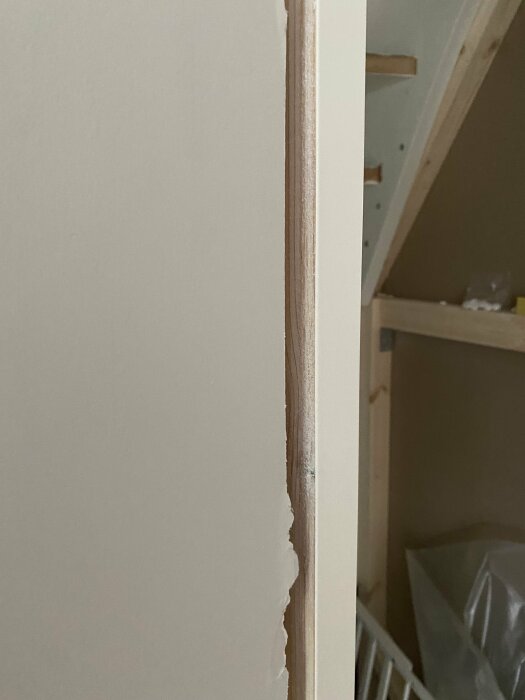 Vägg med skadad kant och synlig plywood, oavslutade byggarbeten i bakgrunden, inomhus renoveringsprojekt.