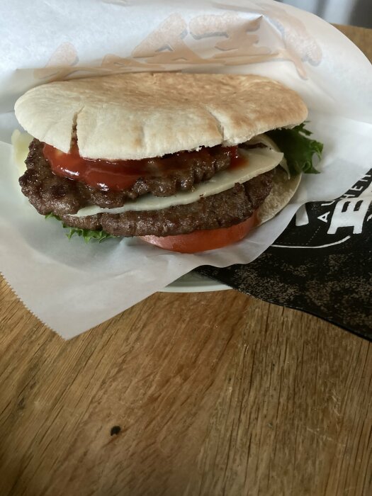 En hamburgare med sallad, tomat, ost, och två köttbiffar i ett pittabröd på ett träbord.