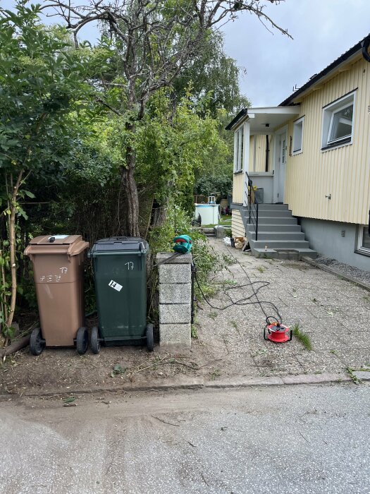 Svenskt bostadshus med sophantering, trädgårdsarbete pågår med hjälp av en grästrimmer, molnig dag.