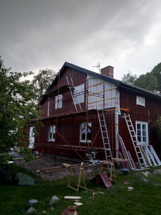 Rött hus under renovering, ställningar, verktyg, grå himmel, obearbetad trädgård, byggmaterial på marken.