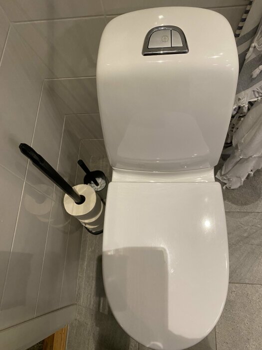 Vitt toalettstol nära grå kakelvägg med toalettpappershållare och extrarullar.