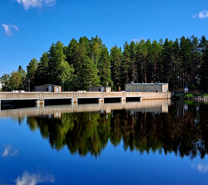 En damm med spegelblank yta, skogsbakgrund under klarblå himmel, arkitektur reflekteras i vatten.