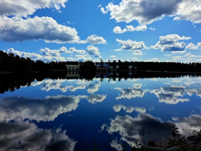 Ett lugnt sjölandskap med spegelblank vattenyta som reflekterar fluffiga moln och kustlinjens byggnader och träd.