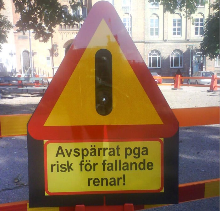 Gul och röd varningskylt med texten "Avspärrat pga risk för fallande renar!" i stadsomgivning.