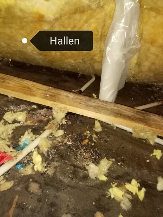 Oisolerat utrymme med skräp, isoleringsmaterial, rör och träreglar. Skylt märkt "Hallen". Oordning och smutsigt.