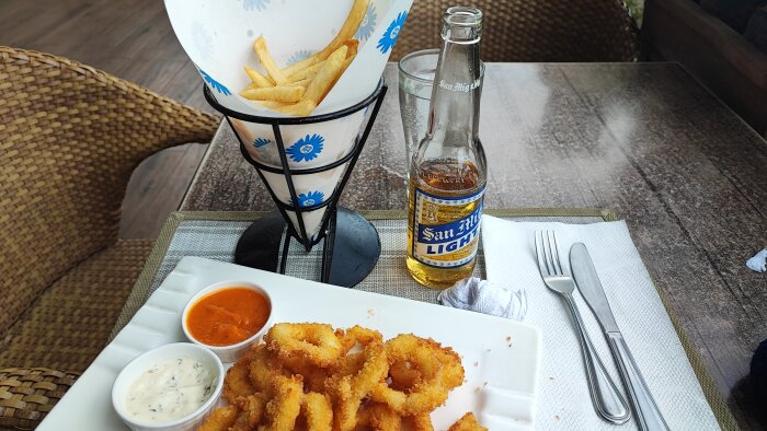 Matplats utomhus med pommes frites i ställ, halvfull ölflaska, friterade calamari och två såser.