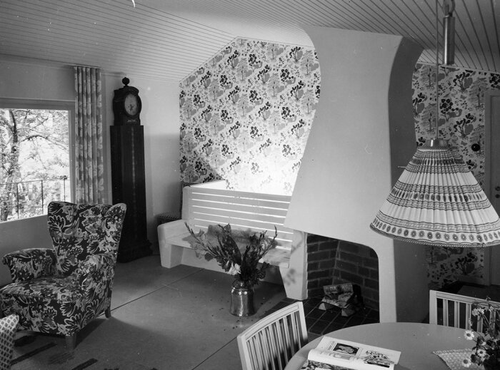 Svartvit bild av ett retro vardagsrum, blommiga mönster, öppen spis, gammaldags inredning och naturutsikt.