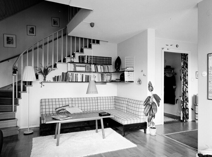 Svartvit bild av ett stilrent vardagsrum med trappa, soffa, bokhylla, matta och dekorativa föremål.