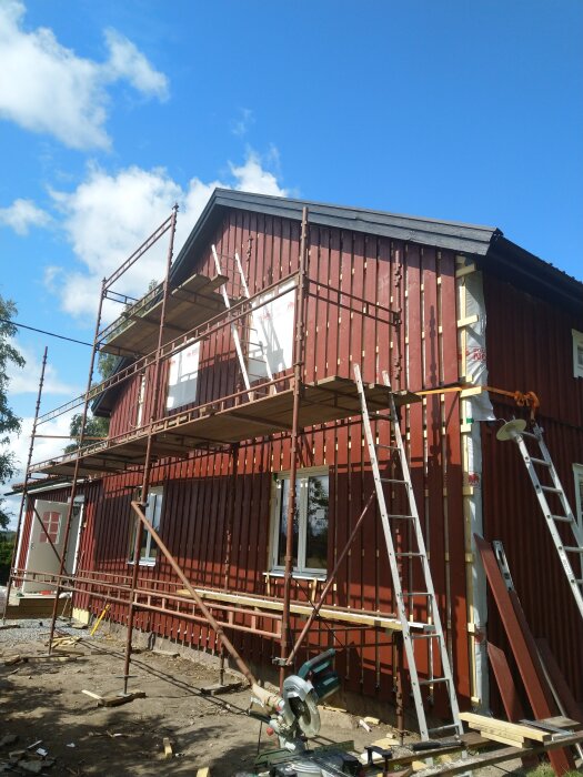 Tvåvåningshus med rött trä, byggställningar, såg och verktyg, renovering pågår, molnig himmel.