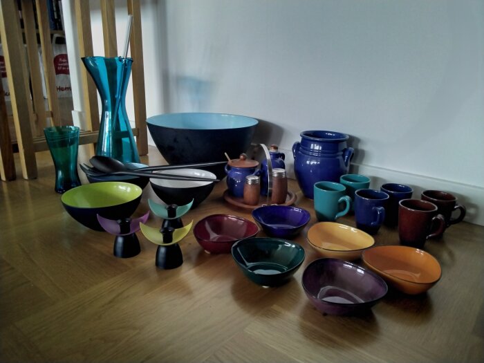Färgglad keramikservis ordnad på trägolv med skålar, koppar och en kanna.