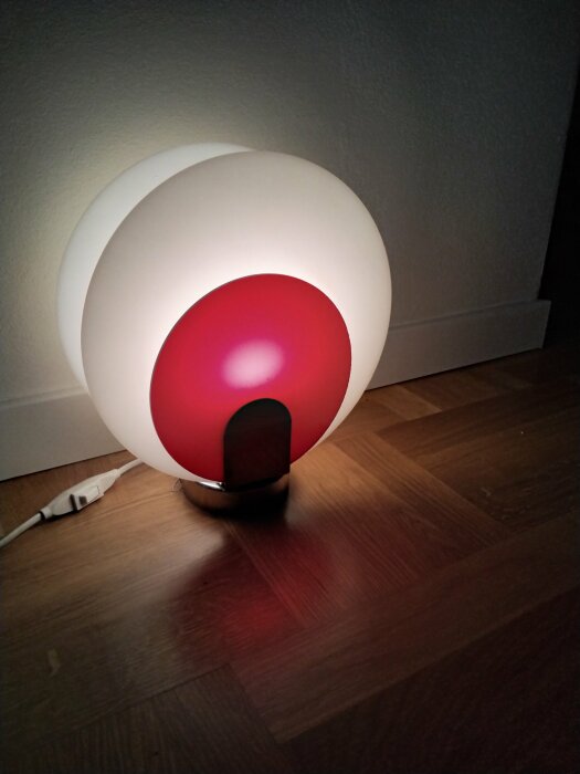 En modern lampa med rund design lyser rött på ett mörkt trägolv nära en vägg.
