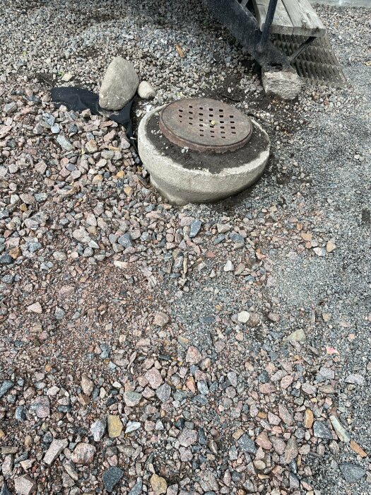 En brunnlock av metall på betongring omsluten av grus och sten, med en metallställning i bakgrunden.