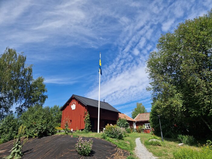 Traditionellt svenskt rött hus, blå himmel, svenska flaggan, grönska, fridfull landsbygdsmiljö.