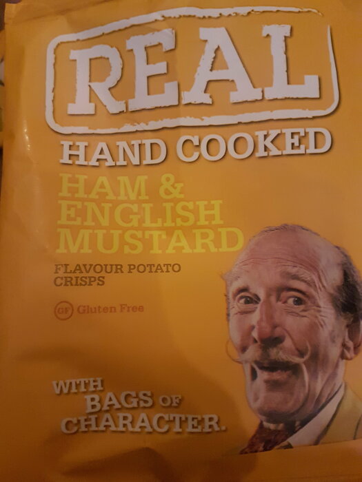 Chipsförpackning, "REAL hand cooked", skinka & engelsk senapssmak, glutenfri, leende man, reklamkampanj, gul bakgrund.