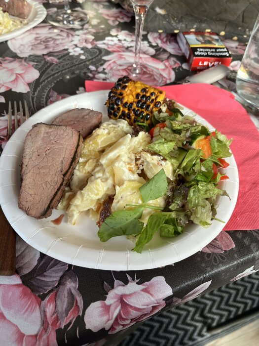 Måltid med kött, sallad, pasta, rostad majskolv, på blommig duk, tom glas, chokladask i bakgrunden.