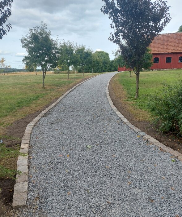 Grusgång kantad av sten, träd, gräsmatta, molnig himmel, del av röd byggnad synlig.