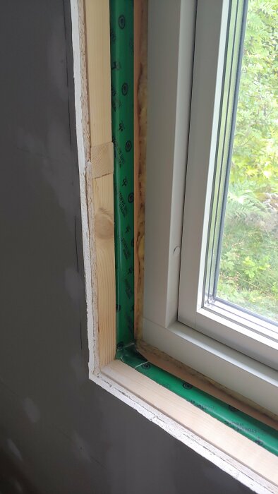 Ett fönster under installation med exponerad isolering, tätningstejp och trästomme mot en vägg.