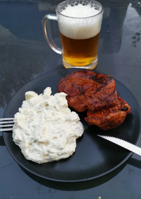 Ett mål mat med grillad köttsteak och potatissallad, serverat med öl, på en svart tallrik.