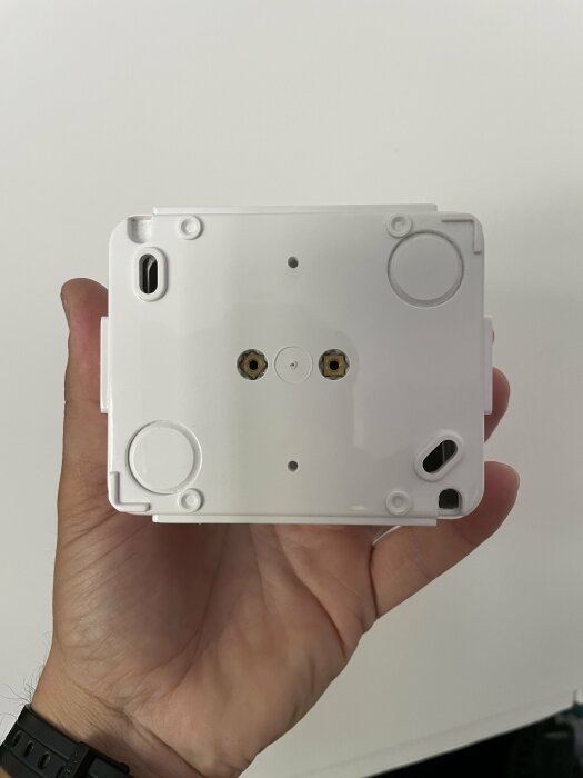 En hand håller vit plastdel, troligtvis till elektronik, med synliga skruvhål och kontaktuttag.