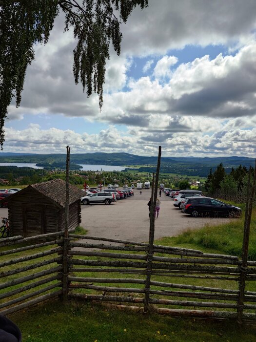 Landskap med parkering, sjövy, molnig himmel, träd, staket, och en person som promenerar.