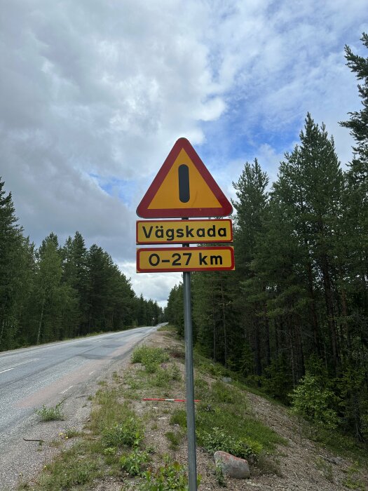 Varningsskylt vid väg, "Vägskada 0-27 km", skog, molnig himmel, asfaltsväg.