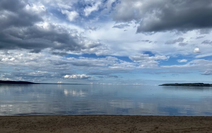 Molnig himmel speglas i lugnt vatten vid strandkant, nära horisont, avlägsna landmassor synliga.