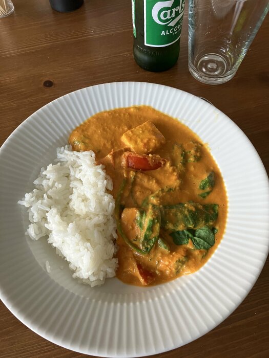 En tallrik med ris och krämig curry, tomater, spenat, en flaska, och ett glas på ett träbord.