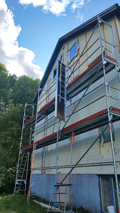 Hus under renovering med byggnadsställningar, stege och oavslutad fasad mot en himmel med moln.