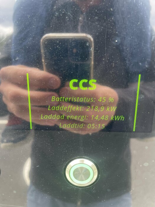 En laddstationsskärm visar batteristatus och laddningsinformation, reflektion av en person som fotograferar skärmen.