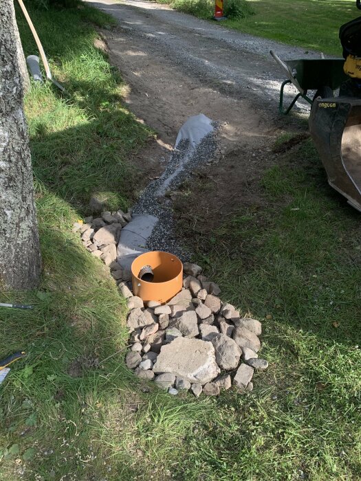 Dräneringsrör och grus vid grävd gräsmarksgrav, omgiven av stenar, skottkärra och trädgårdsverktyg i bakgrunden.