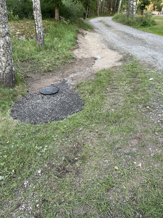 Vägarbete; ny gruslagd yta kring brunnslock bredvid grusväg och träd.