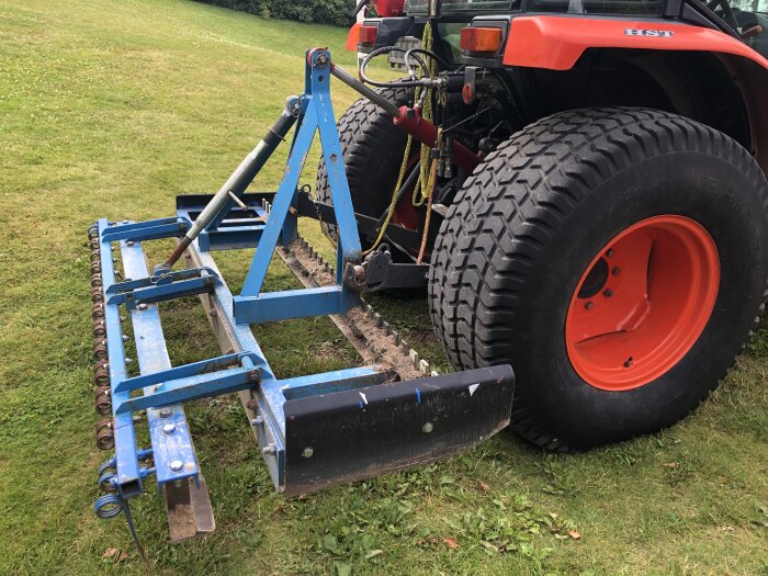 Traktor med gräsplaneringsredskap på grönt gräs, däck synliga, mekaniskt verktyg, grönområde, blå markberedare.