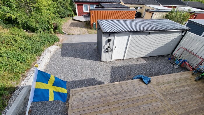 Svensk flagga, träaltan, grusuppfart, skjul, flerfamiljshus, grönska, soligt väder, barnleksaker ute, tyst bostadsområde.