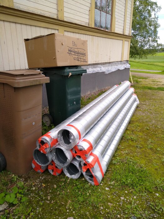 Ventilationsrör staplade utomhus bredvid sopkärl vid gul byggnad med grönska.